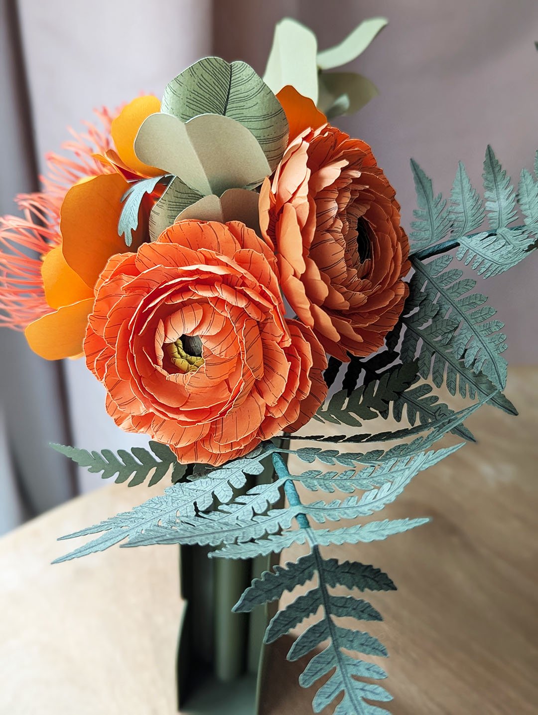 Gros plan sur deux renoncules Clémentine en papier d'art et une fougère vert océan du bouquet Théodore