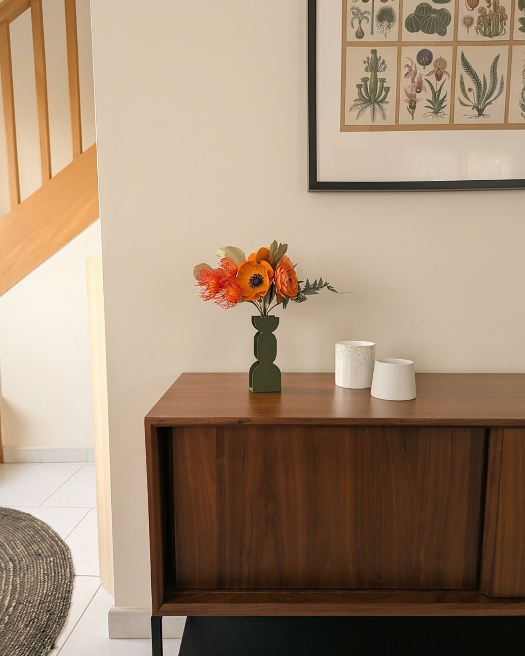 Bouquet de fleurs en papier orange dans un vase vert posé sur un buffet en noyer, à côté de deux pots en céramique blanche